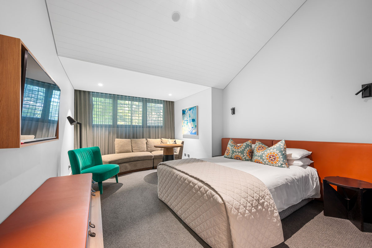 Modern hotel room with queen bed, colourful cushions, sofa, armchair, and TV