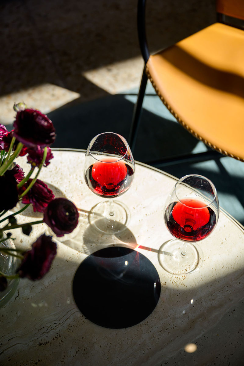 Two glasses of red wine on a marble table with flowers and a yellow chair.