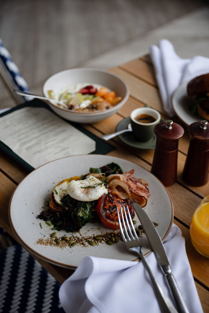 Breakfast plate with poached eggs, grilled tomato, bacon, sautéed greens, coffee cup, juice, and fruit bowl on wooden table.