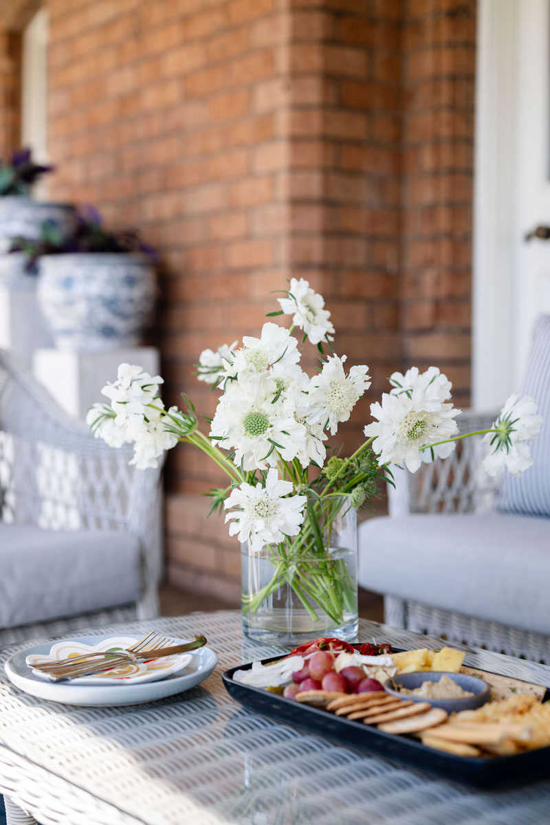 White flowers in glass vase on wicker table with food platter and plates
