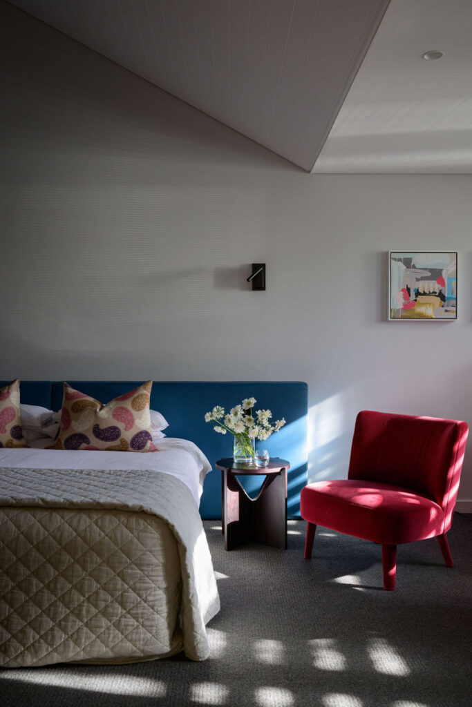 Cozy bedroom with bed, blue headboard, floral pillows, side table with flowers, and red armchair.