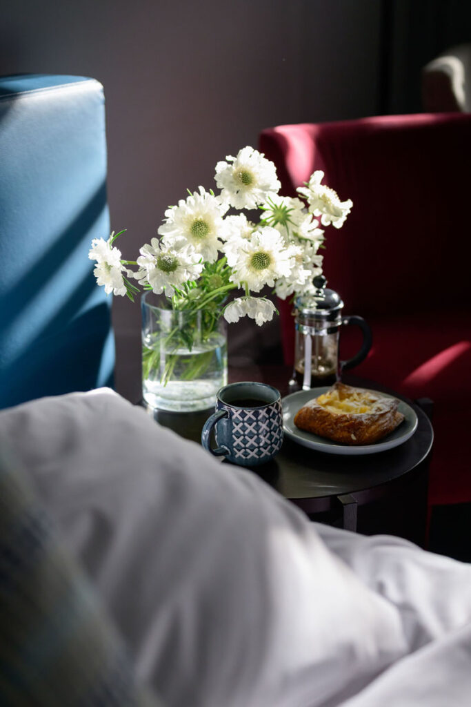 Table with coffee mug, pastry, French press, and white flowers in vase between armchairs