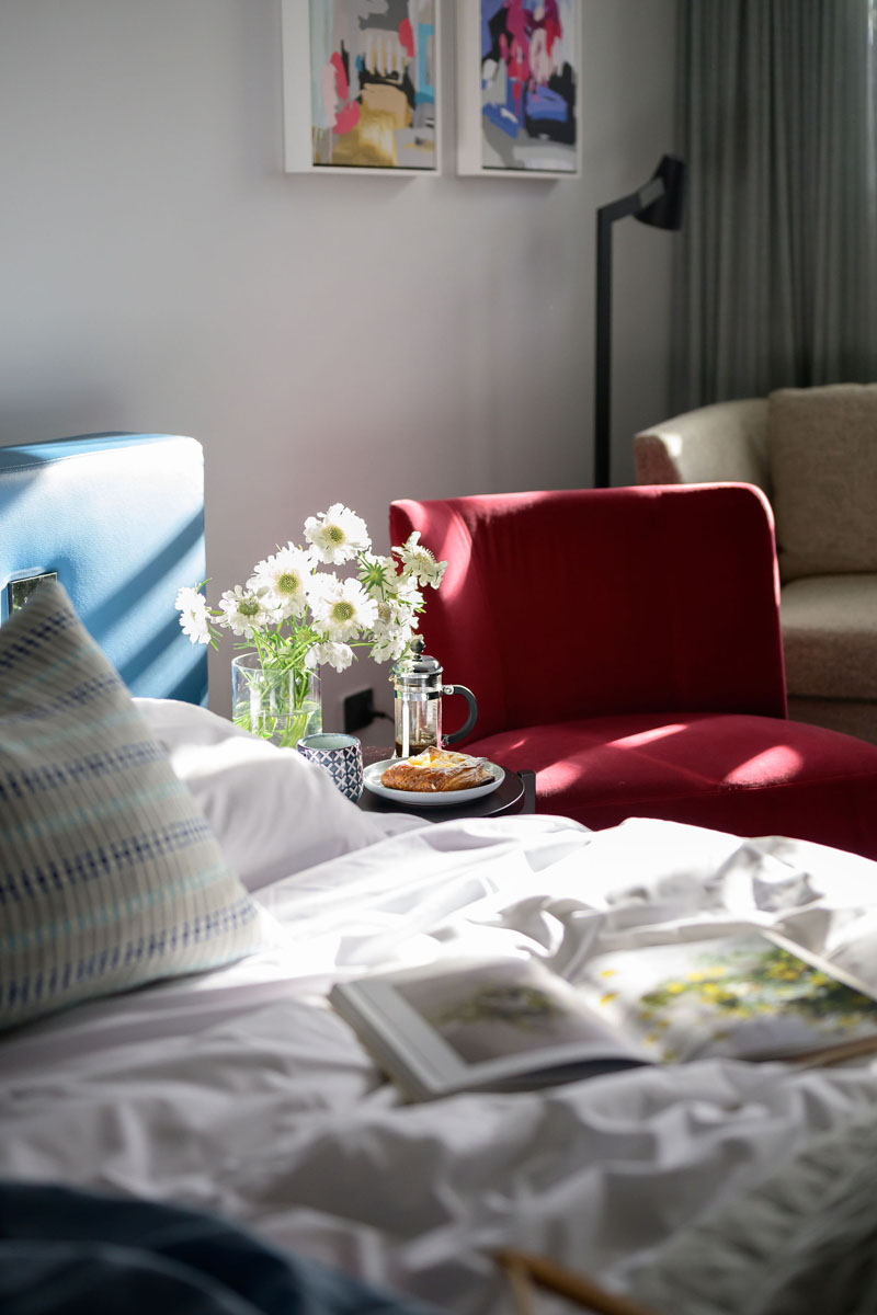 Sunlit bedroom nook with red armchair, white flowers, and open book on bed